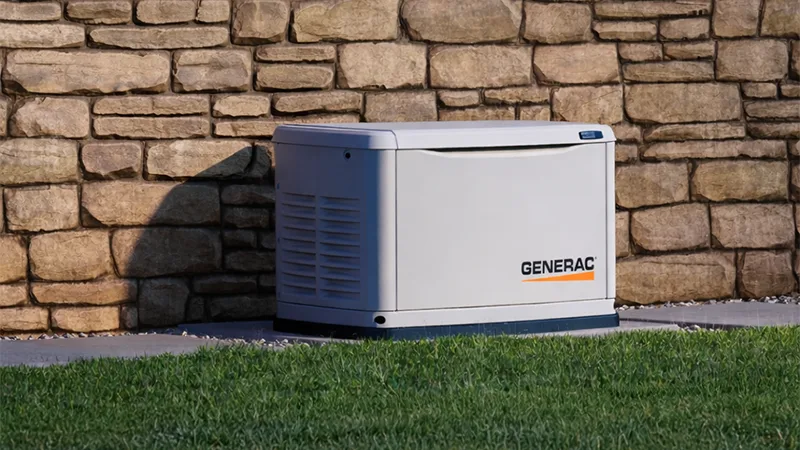 Outdoor Generac standby generator installed on a concrete pad beside a stone wall, with grass in the foreground and natural daylight.