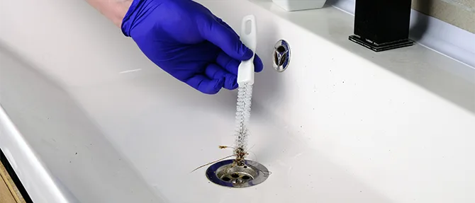 Gloved hand using a drain-cleaning tool to remove hair and debris from a bathroom sink drain.