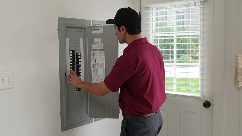 Technician adjusting a residential electrical breaker panel near a door with a window, wearing a maroon uniform and cap.