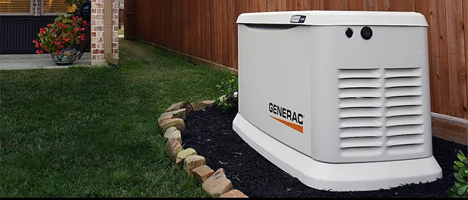 Generac standby generator installed in a landscaped residential yard beside a wooden fence, with grass, stone edging, and garden plants nearby.
