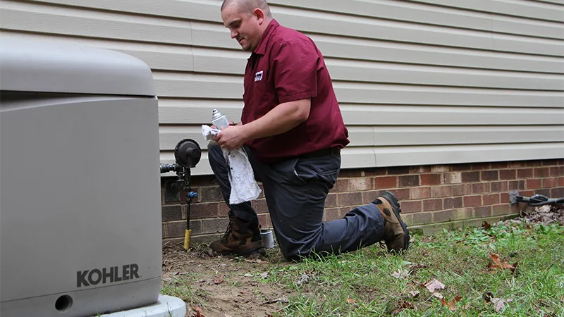 Technician performing maintenance on a Kohler standby generator beside a residential home, kneeling near the gas line connection.