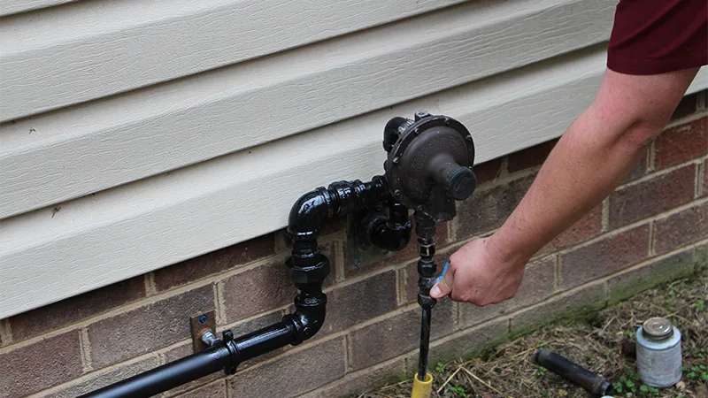Technician adjusting a residential gas line regulator and valve connection mounted on an exterior brick wall.