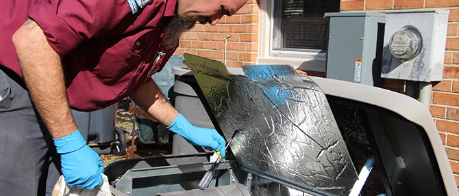 Technician wearing gloves performing maintenance on an open standby generator, inspecting and cleaning internal components beside a residential home