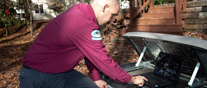Technician servicing an open Generac standby generator using a laptop for diagnostics in a residential backyard with fall foliage.