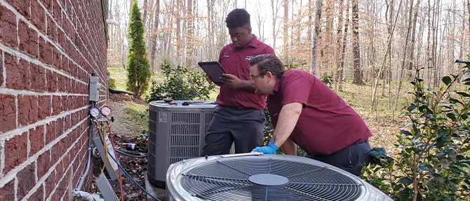 Two HVAC technicians inspecting and servicing an outdoor heat pump unit next to brick house