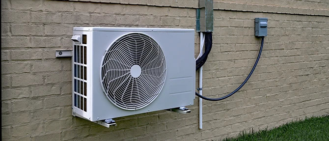 Outdoor condenser unit for a ductless mini-split HVAC system mounted on the exterior wall of a residential home, with electrical disconnect and refrigerant lines professionally installed.