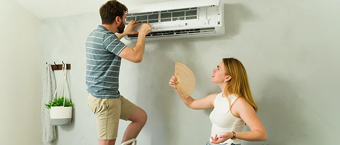 Homeowner inspecting a wall-mounted ductless mini-split air conditioner while a woman cools herself with a hand fan.